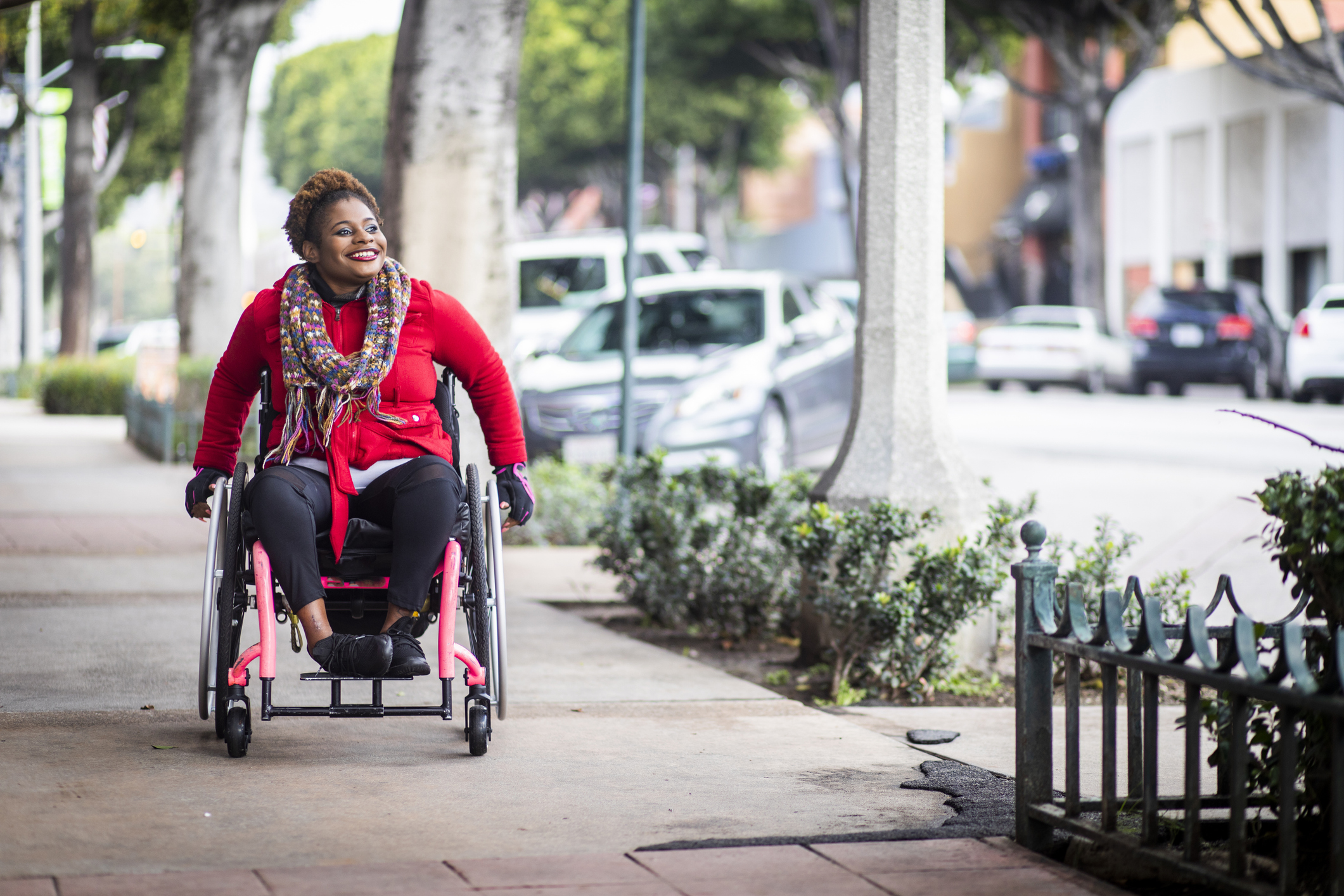 Woman in a wheel chair outside on the sidewalk with a red jacket and a big smile