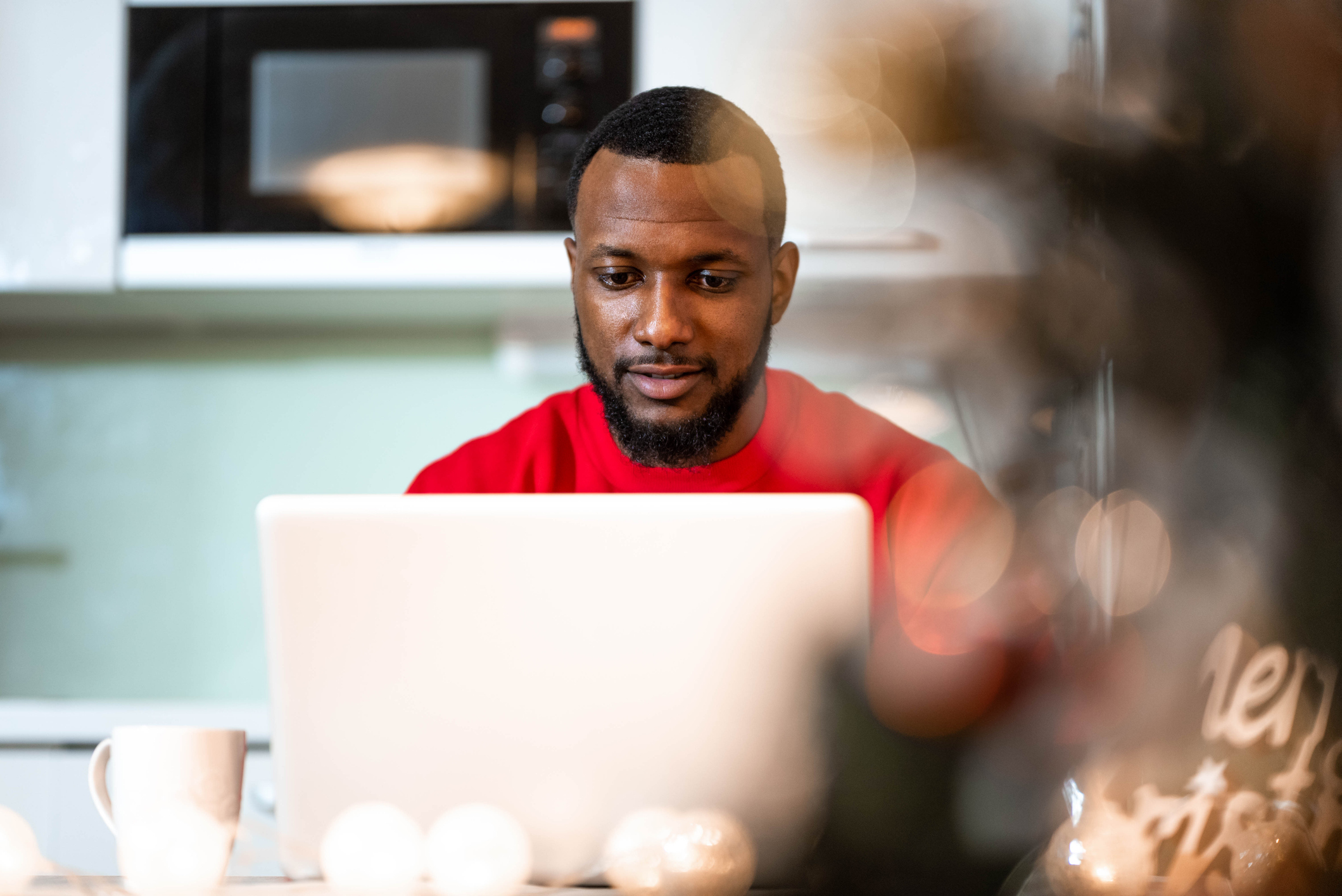 Man in a red shirt, sitting at a table with his laptop and a white coffee cup