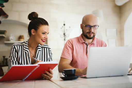 Man and woman sitting at a tiled table, the woman holding a red notepad and the man sitting behind the laptop