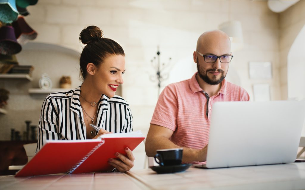 Man and woman sitting at a tiled table, the woman holding a red notepad and the man sitting behind the laptop