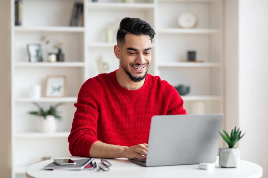 Cheerful attractive businessman in a red sweater sitting at a small table working on a laptop