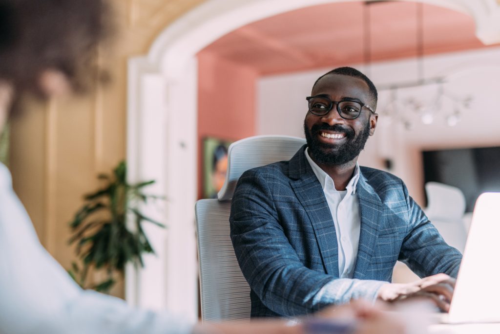 Man with black rimmed glasses wearing a blue blazer sitting in a gray chair at a table talking with another associate.