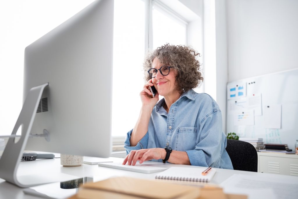 Mature woman wearing denim shirt sitting at the desk in the modern office, using the computer and talking on a mobile phone.