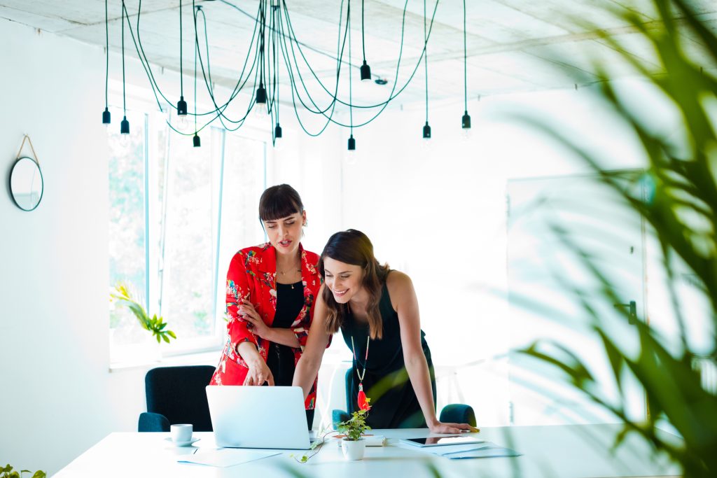 Two women standing at a white desk, looking at a laptop, and string hanging lights above them