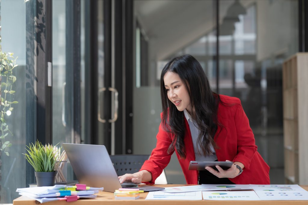 Woman half standing, half leading over a table with graphs, looking at a laptop with a tablet in her hand.
