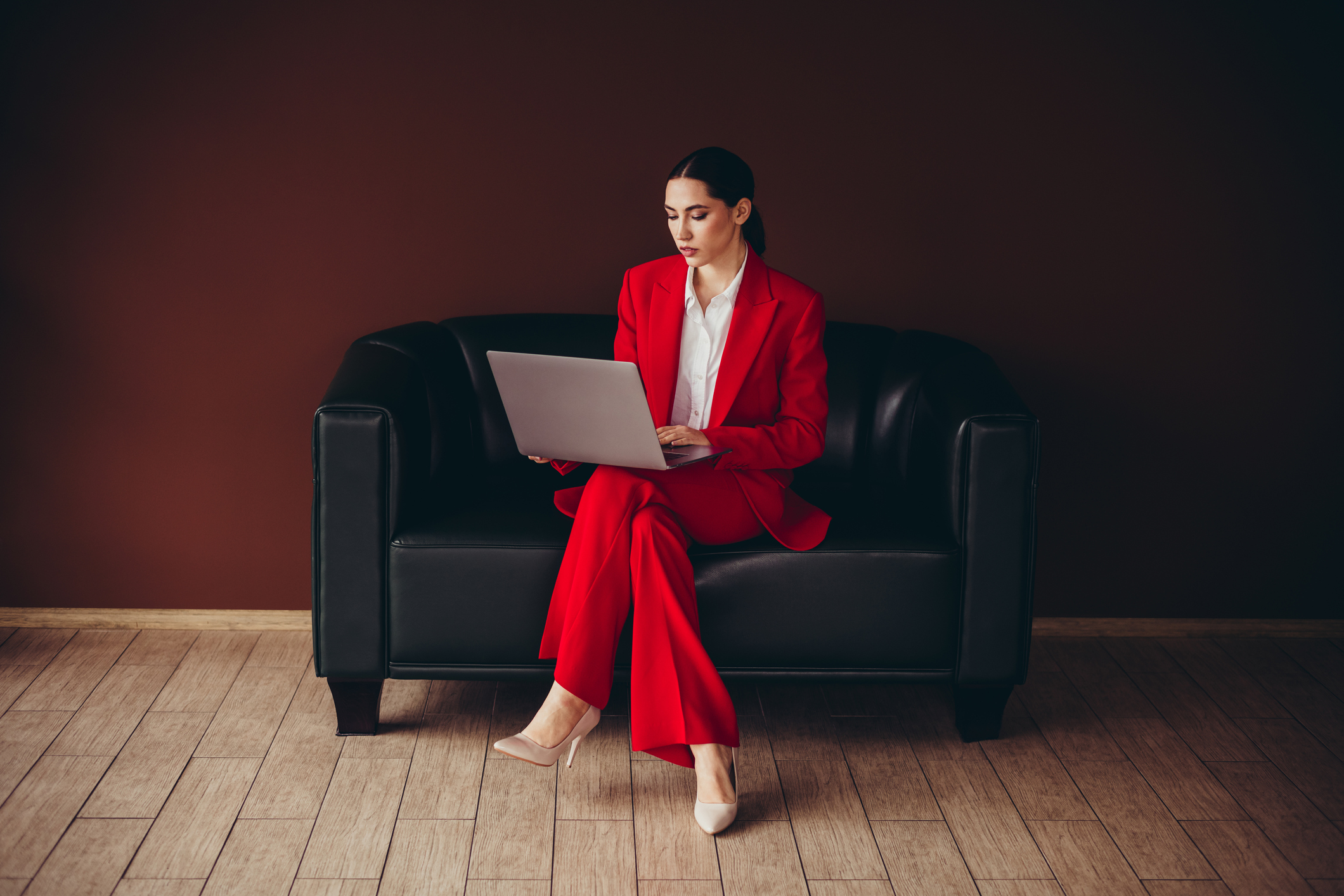 Woman sitting on a black sofa, legs crossed at the knees, red suit and a laptop in her lap