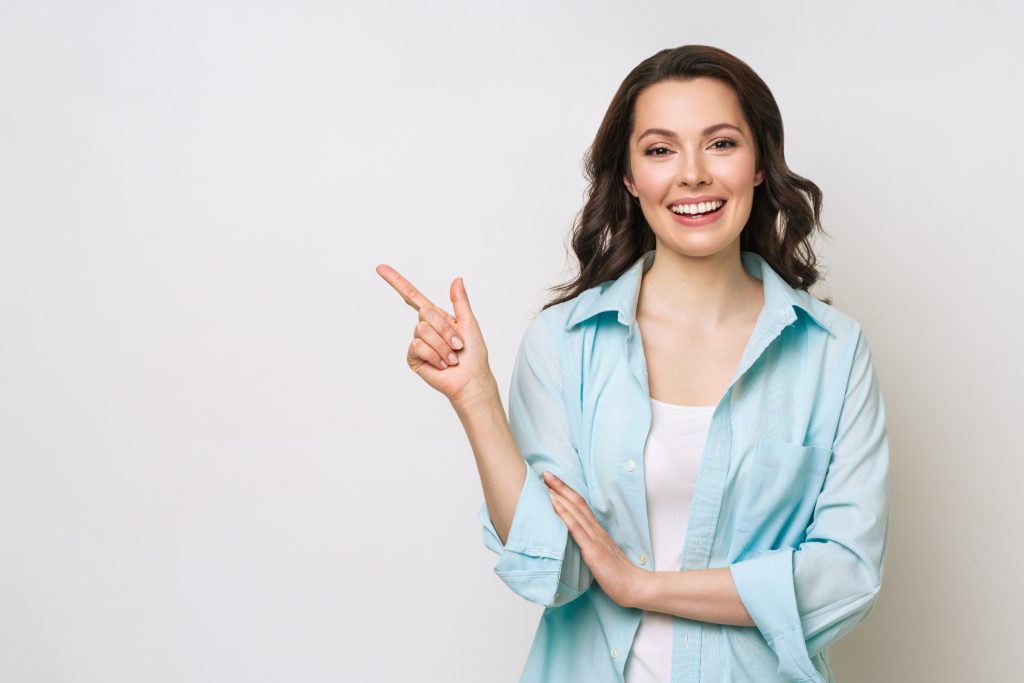Woman with dark brown hair, standing up against a white wall pointing at an empty wall space.