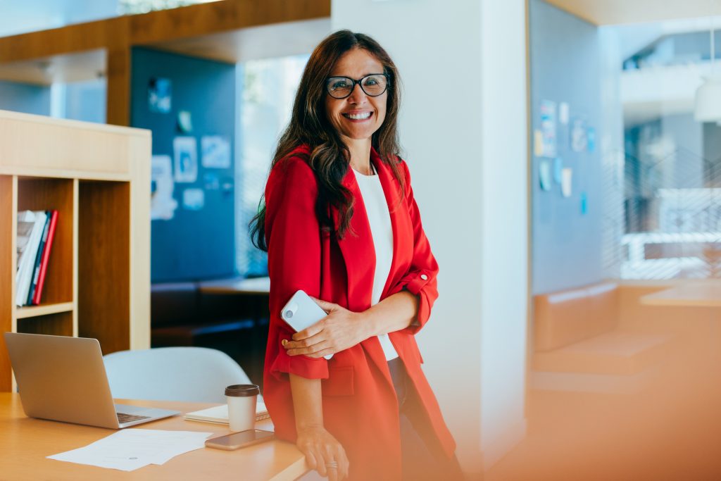 Woman with long hair, wearing red business attire, standing up against a wooden table with her mobile phone in hand