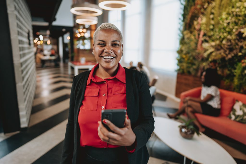 Woman with short hair, wearing a red shirt with a black blazer standing in a room with a mobile phone in her hand