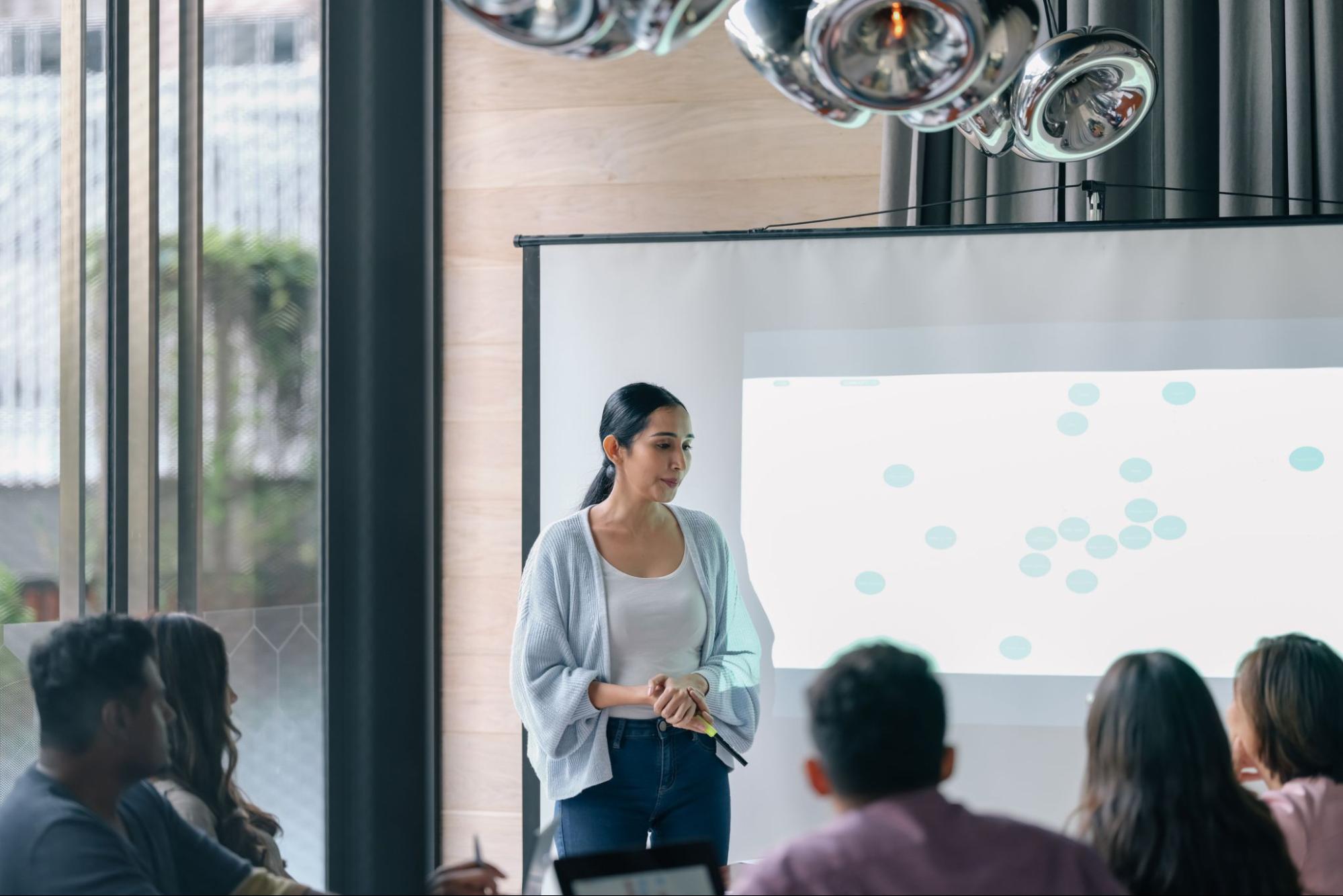 Professional Woman standing up at the front of the room, in front of a projector screen talking to the participants.