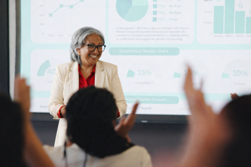 Business woman standing at the front of a room full of other adults with a projected image behind her.